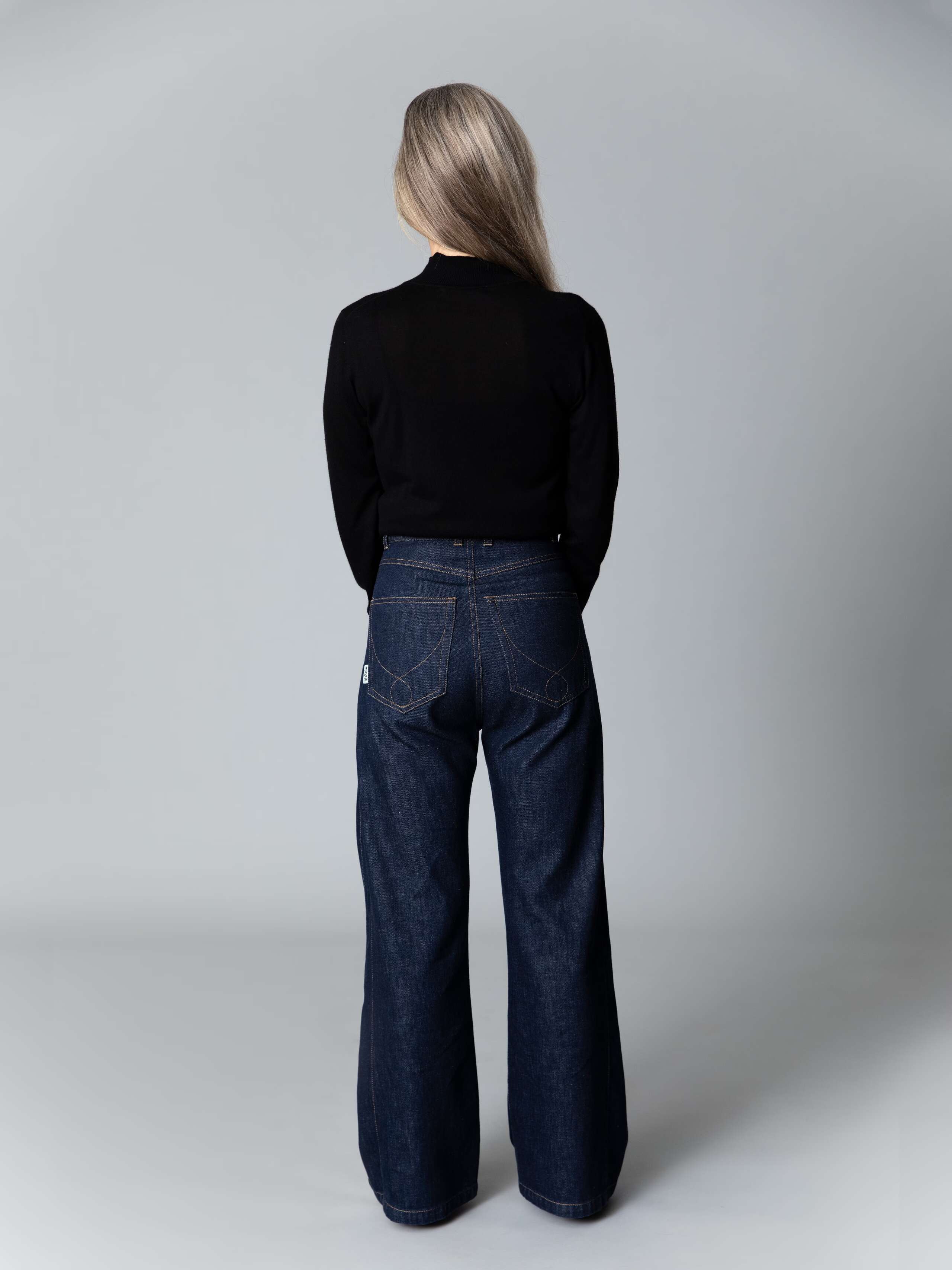 Model with blond hair and light blue eyes wears a black turtleneck pullover and a beuatiful wide leg jeans in dark pure indigo 