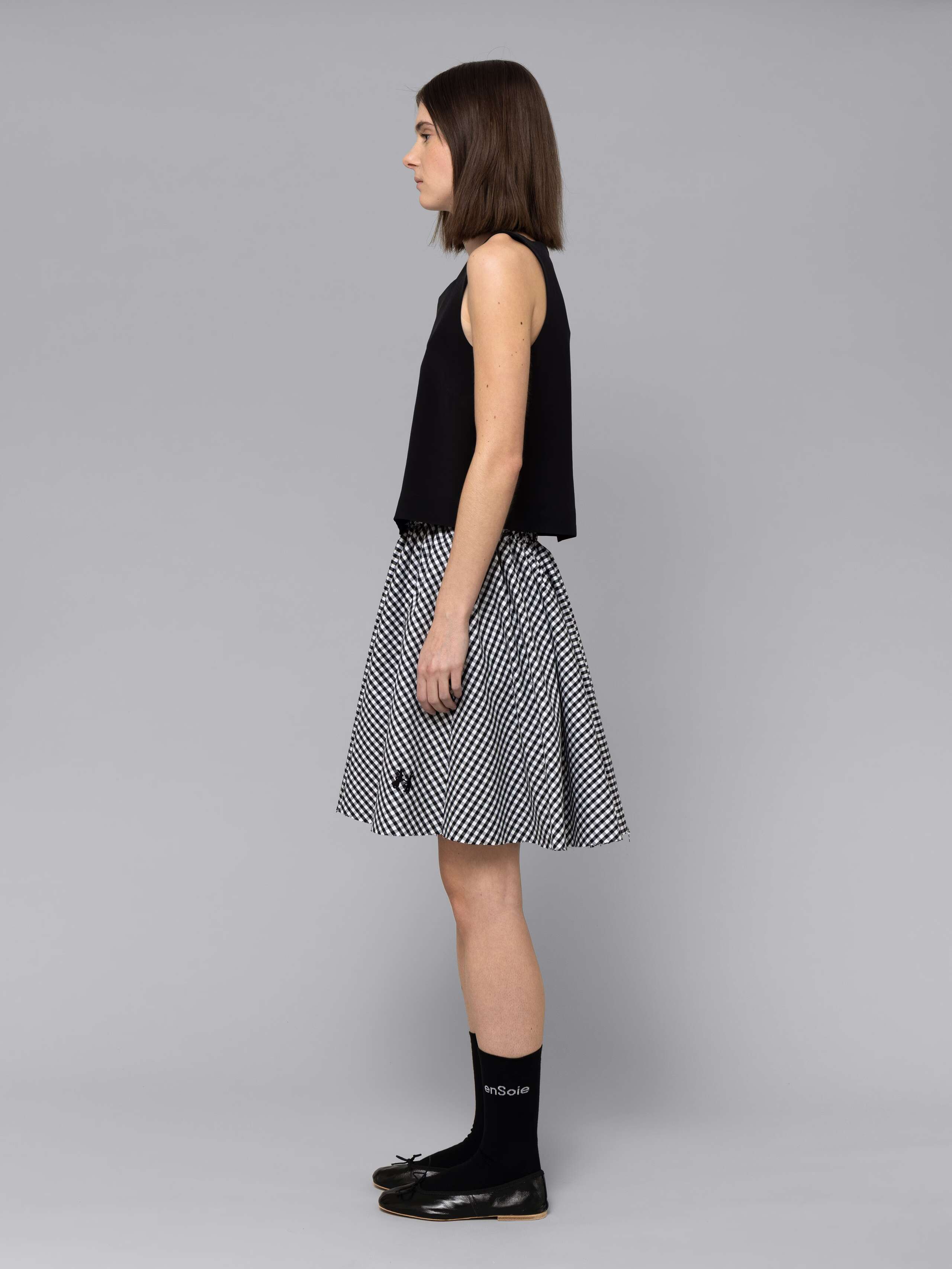 Female model is wearing a combination of a boxy black silk top and a cute short cotton skirt  featuring a beautiful black vichy check pattern