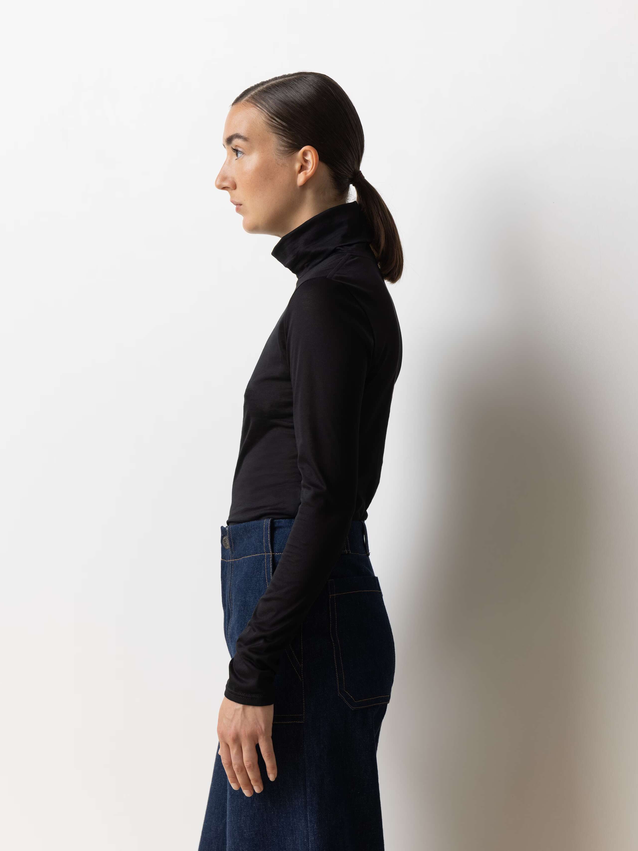 Sideview of a female model wearing a tight fitting turtleneck jumper made of soft black cotton jersey.